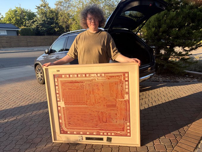 A curly-haired man with glasses stands smiling in a driveway in front of an open car trunk, proudly holding a large framed artwork that displays an orange circuit-board-like design on a cream background. The caption "Subnet 95 - Actual Computer" visible at the bottom of the artwork connects him to the Bittensor Subnet 95 project, literally showing off the "Actual Computer" persona with a piece resembling a computer chip design.