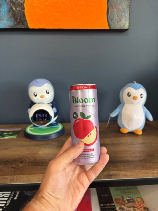 A hand holds up a tall can of "Bloom sparkling energy" in crisp apple flavor in front of a wooden shelf displaying two penguin figurines and a digital clock showing 09:33.