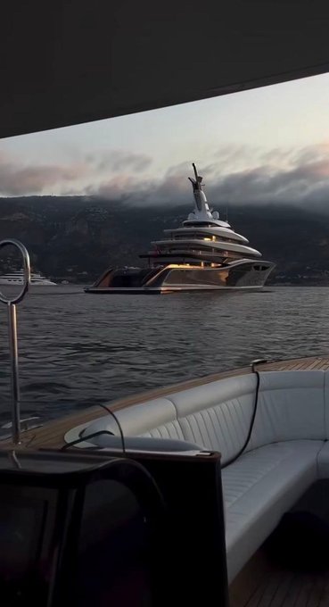 A scenic view from aboard a yacht shows a large luxury cruise ship anchored in the water against a backdrop of hills and a cloudy sunset sky.