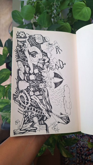 An open sketchbook displaying an intricate black ink doodle/illustration filled with abstract shapes, flowers, a face, and geometric patterns. The sketchbook rests against a backdrop of green potted plants.