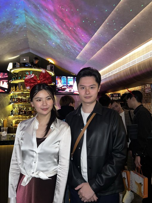 A woman wearing red Minnie Mouse ears and a white satin blouse stands beside a man in a black leather jacket at a vibrant venue featuring a colorful galaxy-projected ceiling and a stocked bar in the background. The image captures the meeting referenced in the post, though neither person pictured appears to match the caption's description of meeting the Bitget CEO Gracy Chen in Dubai and Taipei, with the $389 hack figure not visible in the image itself.