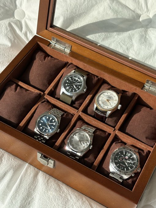 A wooden watch box with a glass lid displays five luxury timepieces on brown cushions, including what appears to be a Rolex Explorer, an Omega with a blue dial, and a GMT-style watch. The collector is looking to expand the collection with a Cartier or Bulgari next year, hoping to fund the purchase with gains from an upcoming crypto bull market.