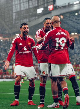 Manchester United football players celebrate together on the pitch; “HEUNME” and the number 19 are visible on the back of one player’s red jersey.