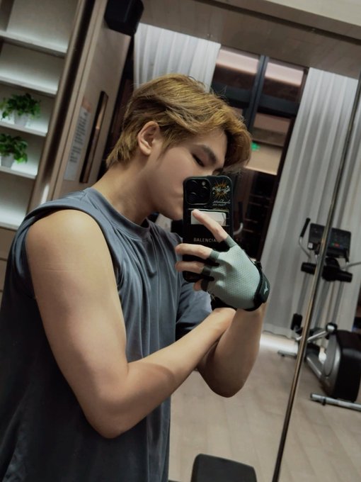 A mirror selfie of a young man with dyed hair wearing a grey sleeveless shirt and a workout glove, holding a phone with a cardholder case in a gym setting.