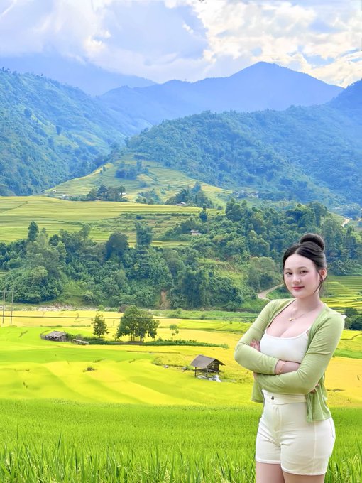 A young woman in a green cardigan and white shorts stands with crossed arms in front of lush green rice terraces and mountains in the background.