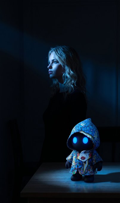 A woman with wavy blonde hair stands in profile against a dark background, gazing thoughtfully to the side, while a small hooded figurine with glowing blue eyes and a colorful patterned robe sits on a wooden table beside her. The image showcases the "Myo x Sabet" digital collectible, highlighted in the post as an elegant piece from the artist collaboration with SUYE on VeVe.
