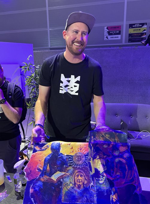 A smiling bearded man wearing a black cap and a black t-shirt with a white '芝' (zhī) Bitcoin-style logo, posing at what appears to be a convention while holding up a colorful Bitcoin-themed artwork.