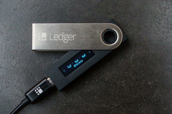 A Ledger hardware wallet with its swivel cover bearing the "Ledger" logo is shown alongside the device displaying "Bitcoin" on its screen, illustrating the type of physical crypto storage device allegedly compromised in the reported $86 million theft from Ledger users who purchased through an unauthorized reseller.