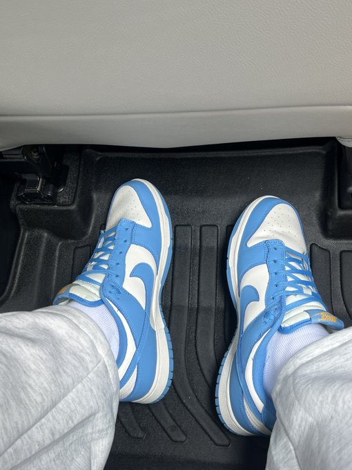 A pair of blue and white Nike Dunk Low sneakers worn with white socks, photographed from above inside a car on a black floor mat.