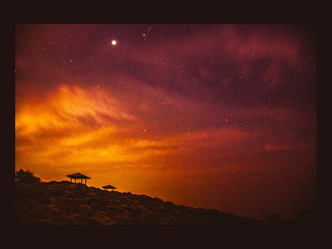 A nighttime landscape photograph showing a silhouetted hill with two small gazebo structures under a dramatic sky featuring the Milky Way, stars, and reddish-orange glowing clouds.