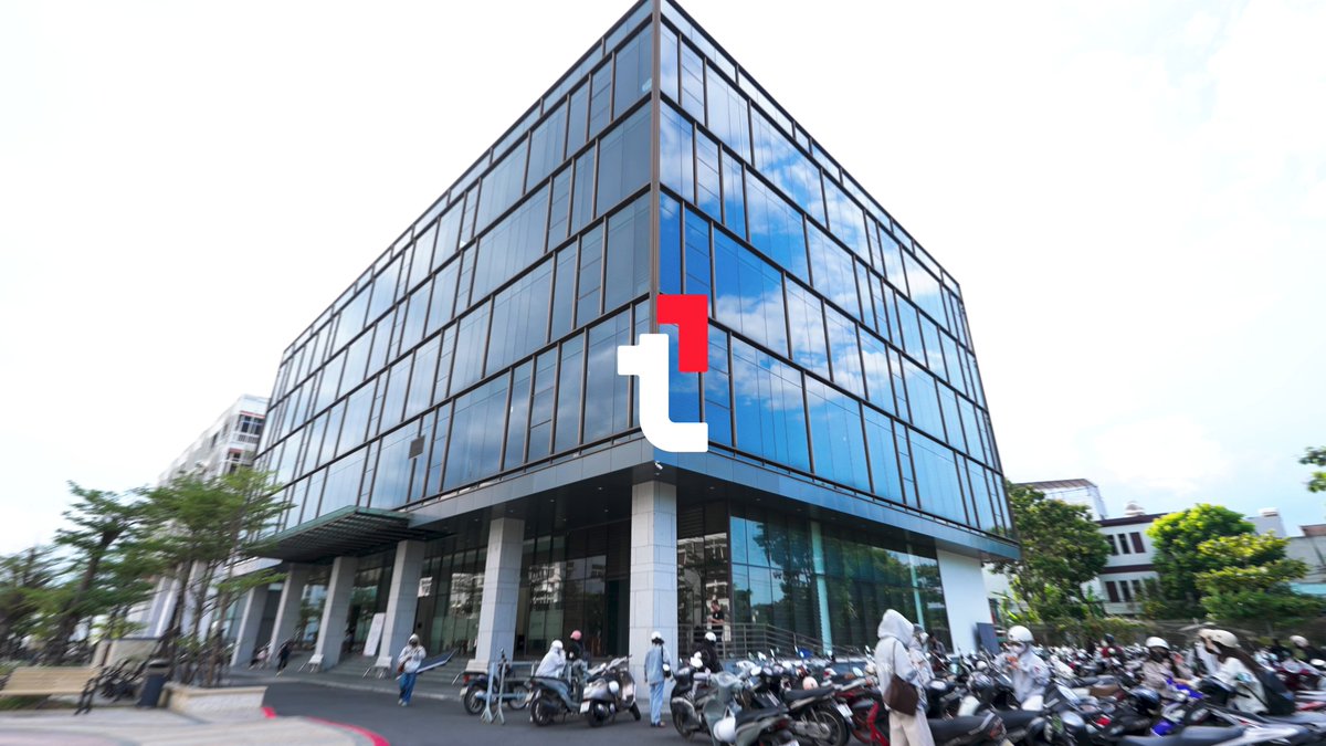 The image shows a large glass building with a red logo on its side, possibly representing a company or organization. There are several motorcycles parked outside of the building, indicating that this could be a popular gathering spot for motorcycle enthusiasts. The photo also includes text about an upcoming event at a university in Vietnam and encourages people to share their experiences by tagging @Team1VN Uni Tour.