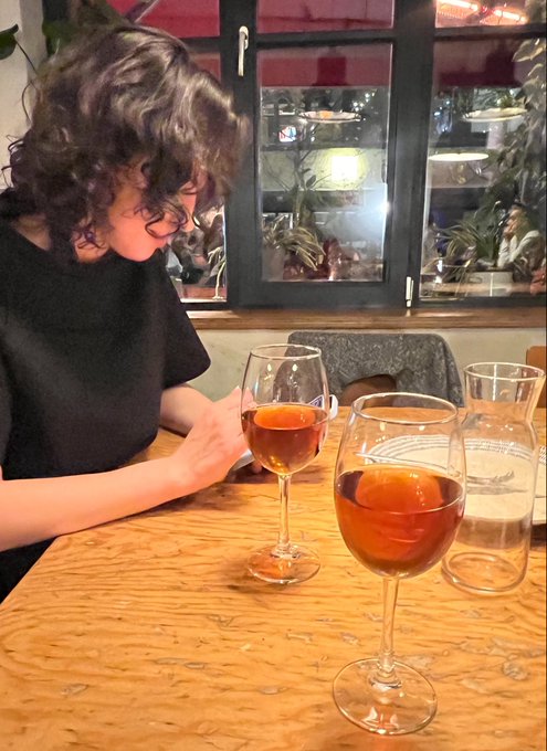 A child with curly hair sits at a wooden table in a restaurant looking at a phone, with two wine glasses containing an amber beverage and a water carafe in the foreground.
