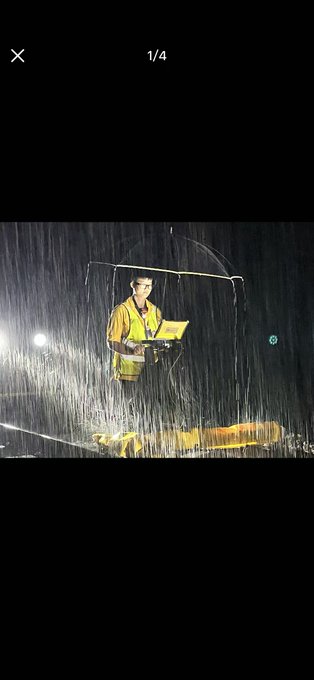 A photo of a person in a yellow shirt holding a clear umbrella in heavy rain at night, standing on a wet surface illuminated by light.