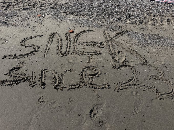 The names 'SANGEK' and 'SHENSHEN' are handwritten together in the wet sand of a beach.