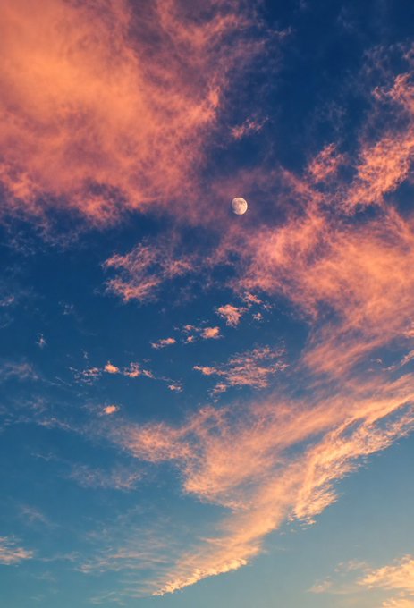 A daytime sky with wispy pink and orange clouds illuminated by sunset light, with a pale gibbous moon visible in the upper portion.