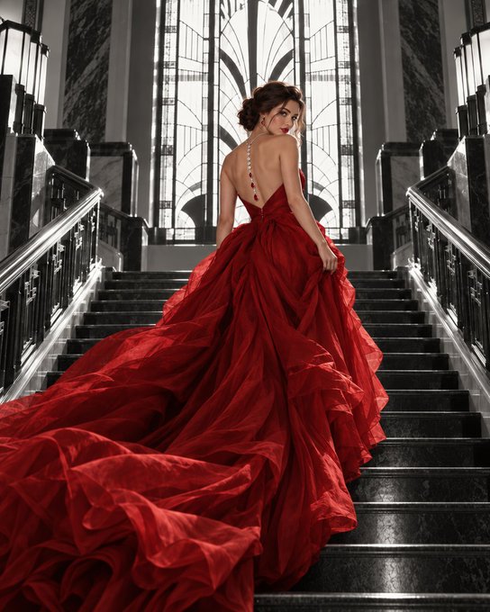 A woman in a voluminous, backless red gown stands on a grand black staircase, the dress's long train trailing dramatically down the steps.