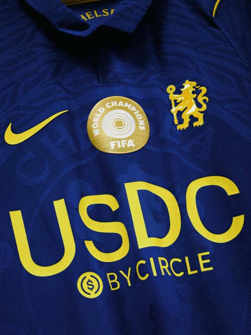 A navy blue Chelsea FC away jersey features a gold "FIFA World Champions" badge, the iconic yellow lion crest, and sponsor "USDC by Circle" in bold lettering across the chest. The "🔵" emoji corresponds to the blue color of the kit, celebrating Chelsea's FIFA Club World Cup victory with a USDC sponsorship tie-in.