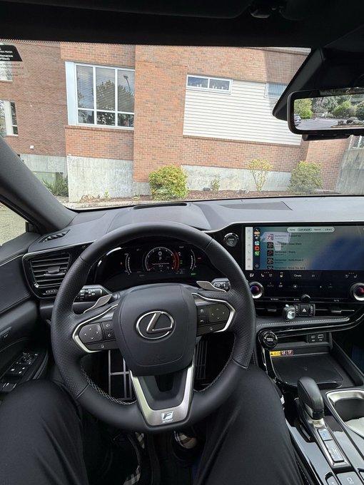 Interior view from the driver's seat of a parked Lexus, showing the steering wheel, dashboard, and infotainment screen with a brick building visible through the windshield.