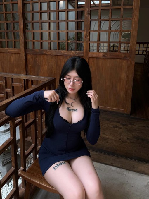A woman with long black hair and glasses sits on a wooden stool, wearing a navy blue zip-up dress that displays a 'Teresa' tattoo design and '1999' on her thigh.