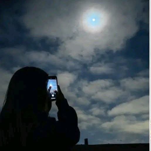 A silhouette of a person holding up a smartphone to photograph or capture the bright sun shining through scattered clouds in a blue sky.