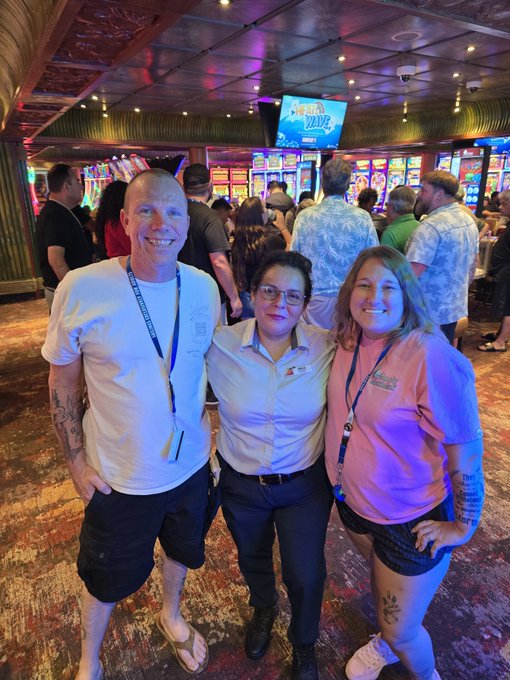 Three smiling people pose together inside a brightly lit casino, with rows of colorful slot machines and a crowd of patrons visible in the background.