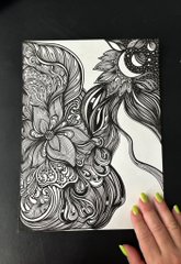 A black and white intricate ink drawing on paper featuring a face intertwined with floral and mandala patterns, held by a hand with neon green nails.
