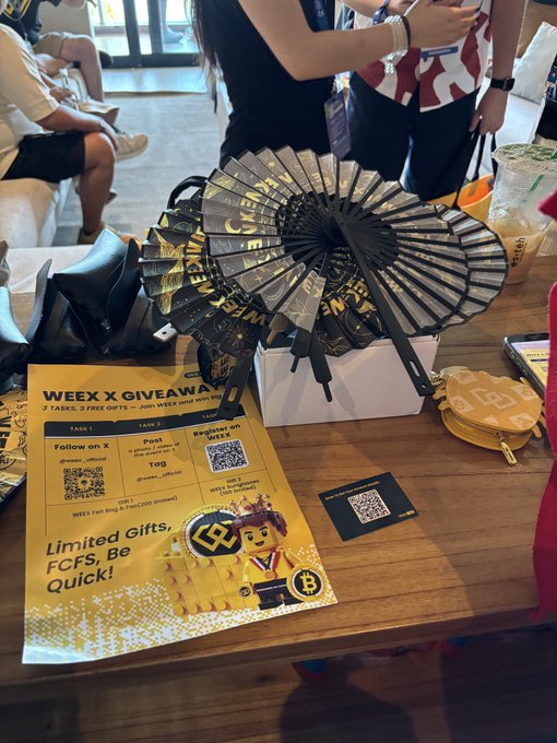 Photo of a WEEX x GIVEAWAY booth featuring folding fans with WEEX branding, a promotional flyer offering 2 tasks and 3 free gifts, and a mascot figure, set at a crypto conference event.