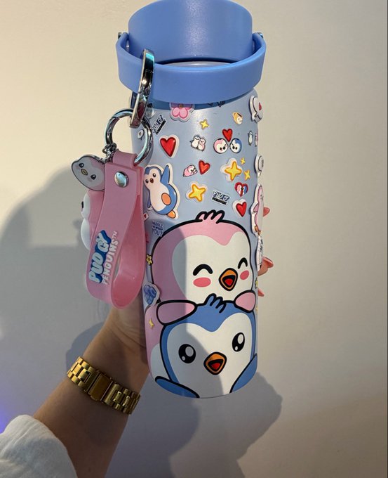 A photograph of a hand holding a light blue insulated water bottle decorated with cartoon penguins and flowers, featuring a blue lid and a pink 'Penguin Adventure' retractable leash strap.