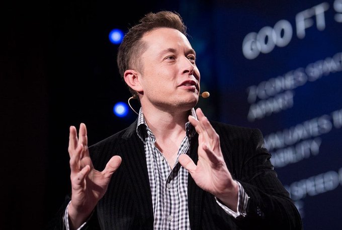 Elon Musk, wearing a checkered shirt and dark blazer with a headset microphone, speaks expressively on stage with both hands raised in a gesturing pose. The backdrop displays partial text reading "GOOF T" and "IT COMES FRO..." alongside bullet points, suggesting a conference setting that ties to the post's theme of Musk's predictions about AI-driven abundance.