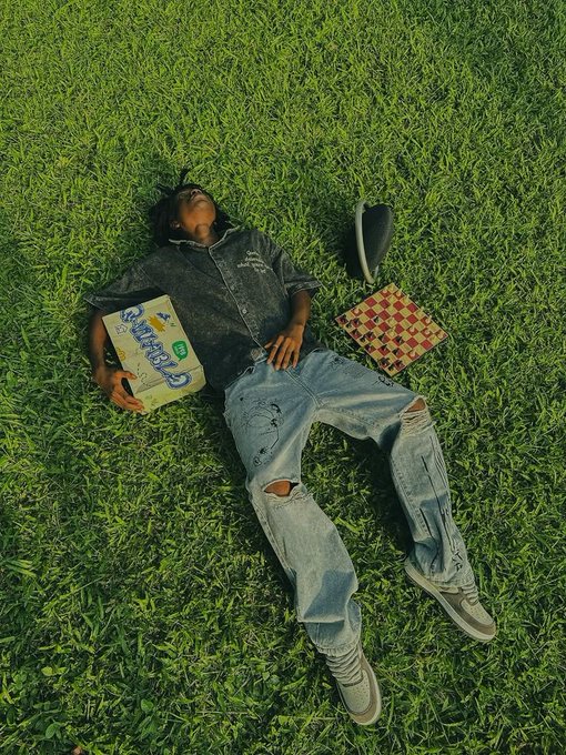 A young person lying on green grass wearing a denim shirt and jeans, holding a Pizza Hut box labeled 'No Strings', with a black bag and a checkered item nearby.