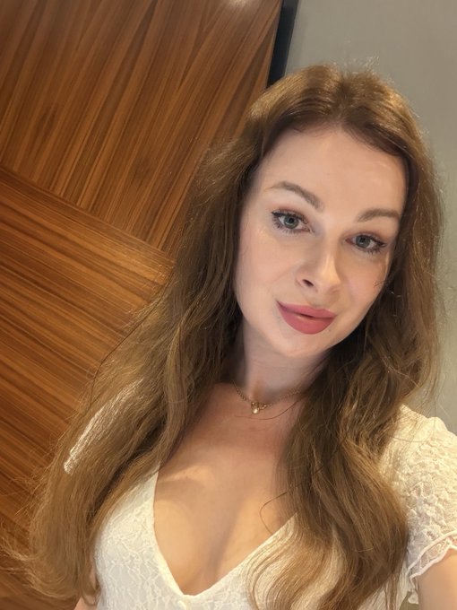 A woman with long brown hair wearing a white lace top, posing for a selfie.