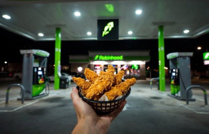 A hand holds a basket of golden chicken fingers in front of a Robinhood gas station at night, with the brand’s green logo illuminated overhead. The scene contrasts the tangible snack—eaten—with the digital “tendies” promised by the platform.