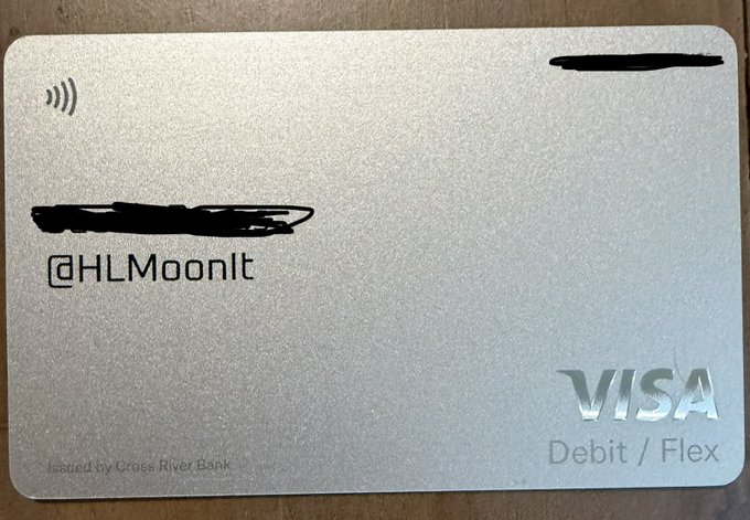A metallic Visa Debit/Flex card issued by Cross River Bank sits on a cardboard surface, featuring a contactless payment symbol in the top-left corner and the personalized handle "@HLMoonIt" displayed on the left side. The card matches the post's excitement about receiving the X Money card, showcasing the custom handle mentioned by the user alongside the Visa branding.