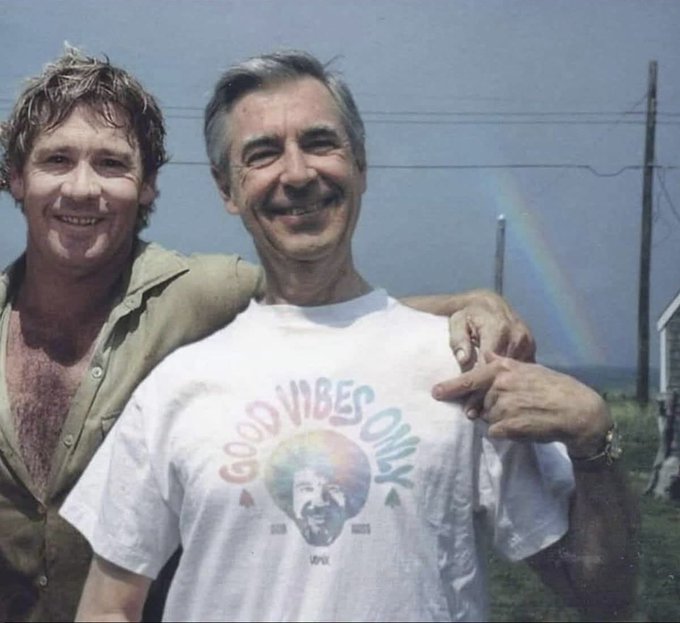 Two men pose together outdoors with a rainbow visible in the background; the man on the right wears a white 'Good Vibes Only' t-shirt featuring a portrait of Fred Rogers.