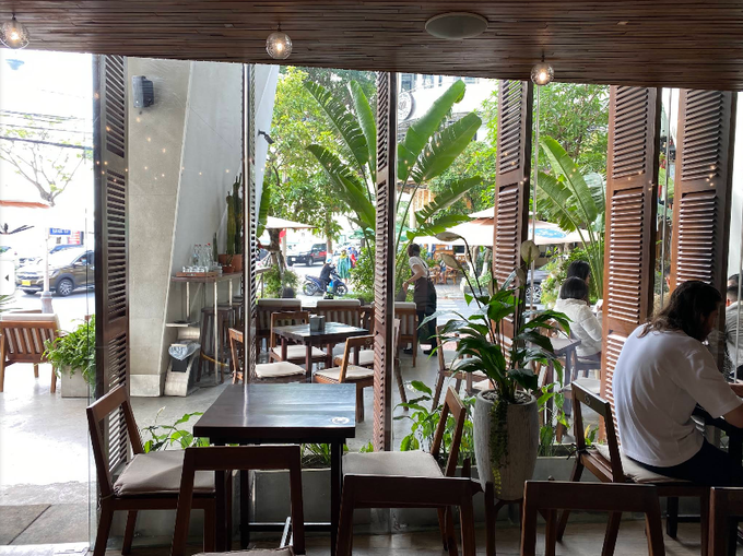 View from inside a café looking out through wooden shutter doors at a tropical street with palm trees and patrons seated at tables.