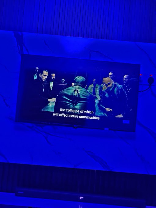 A television screen in a blue-lit room displays a scene of men in suits gathered around a table, with the subtitle 'the collapse of which will affect entire communities'.