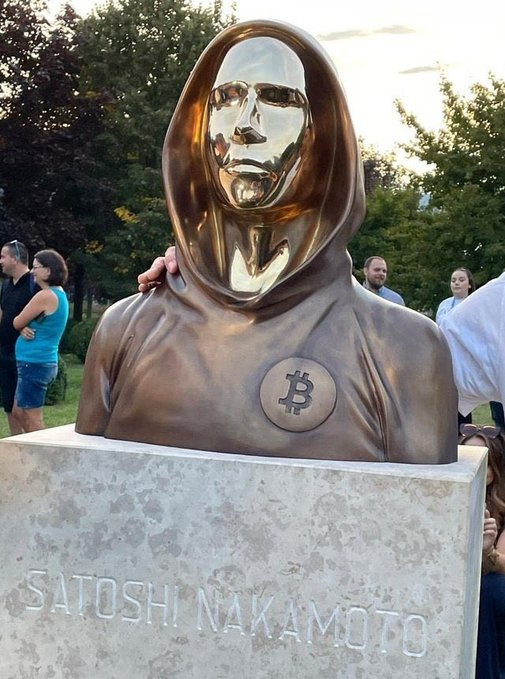 A bronze statue of a hooded figure with a Bitcoin logo on its chest, mounted on a stone pedestal inscribed with 'SATOSHI NAKAMOTO', surrounded by people in a park setting.