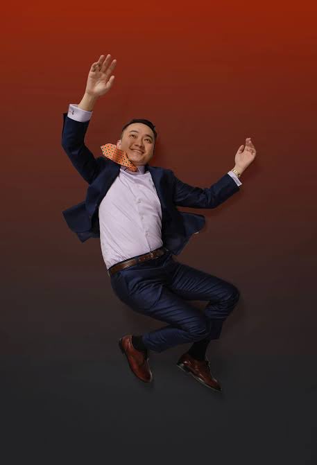A man in a navy suit and orange patterned bow tie is captured mid-jump against a gradient brown background, with his arms and legs spread out.