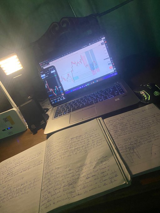 A dimly lit desk with a laptop displaying trading charts, accompanied by a smartphone, notebooks with handwritten notes, and a portable light
