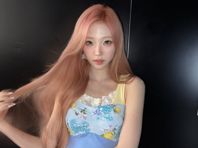A woman with long peachy-pink hair wearing a colorful floral print top poses against a dark background.