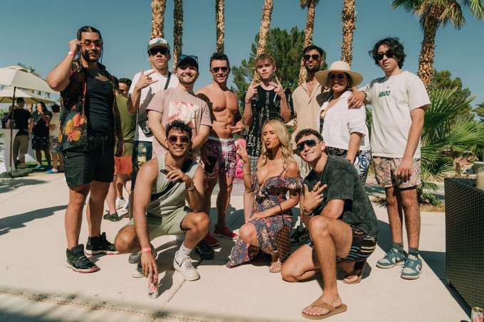 A group of around eleven people pose together outdoors at what appears to be a sunny poolside or beachside location with palm trees in the background, dressed in summer attire.
