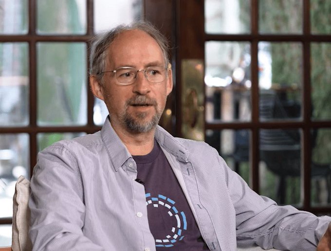 A middle-aged man with glasses and a gray beard, wearing a light blue button-up shirt over a dark t-shirt featuring a blue circular Bitcoin logo, sits in front of a multi-paned window. The image relates to the post as it likely depicts Adam Back, a prominent Bitcoin figure, who is referenced as having a standing buy order for Bitcoin.