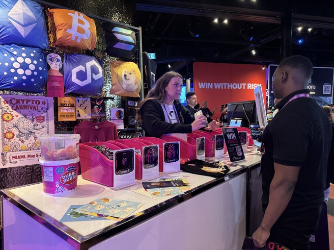 A booth at the Crypto Carnival featuring cryptocurrency-themed merchandise including Bitcoin, Ethereum, and Dogecoin pillows, with staff assisting customers.