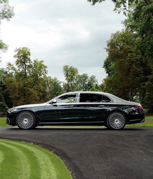 Side profile of a black luxury Mercedes-Maybach sedan parked on an asphalt driveway surrounded by lush green trees and manicured lawn.