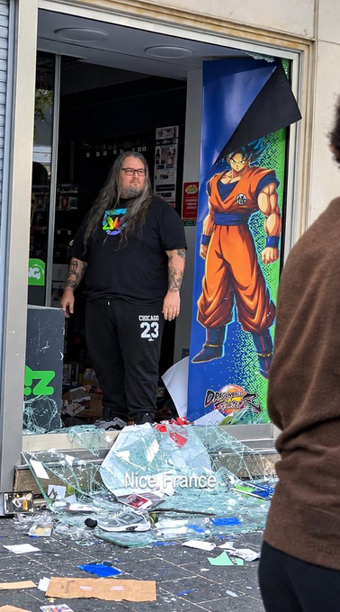 a man with long hair and glasses standing in the doorway of a shop with shattered glass on the floor, next to a dragon ball fighterz banner, with "Nice, France" labeled on the image.