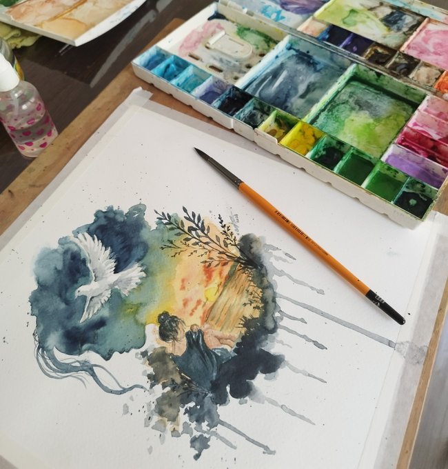A photograph of a watercolor painting setup featuring a painting of a figure with a white bird, alongside a palette of watercolors and an orange paintbrush on a wooden desk.
