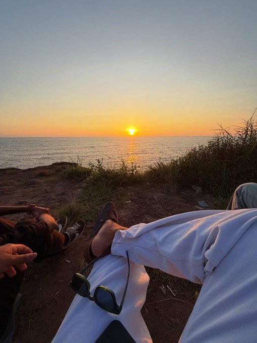 A first-person view of two people relaxing on a grassy cliff at sunset, with the ocean stretching to the horizon. A pair of green sunglasses and white pants are visible in the foreground.