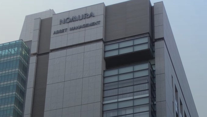 A low-angle exterior shot of a tall, modern office building with the name 'NOMURA ASSET MANAGEMENT' displayed on its upper facade.