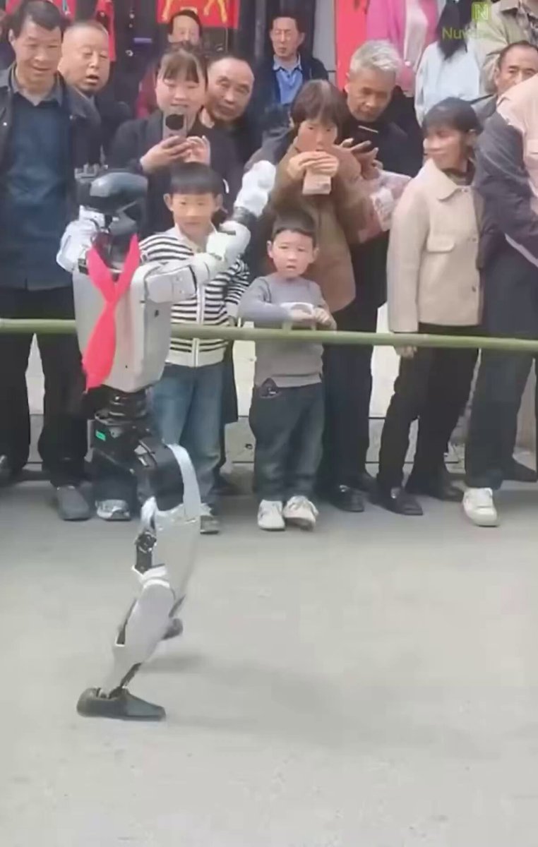 The image features a robot walking down a street, with people watching from behind a railing. There are at least 13 people visible in the scene, some of whom appear to be taking photos or recording videos using their cell phones. The crowd seems interested in the robot's actions and is likely observing its movements closely. The post about Bitcoin appears to be related to this event, as it suggests that there might have been a significant development or news related to cryptocurrency recently.
