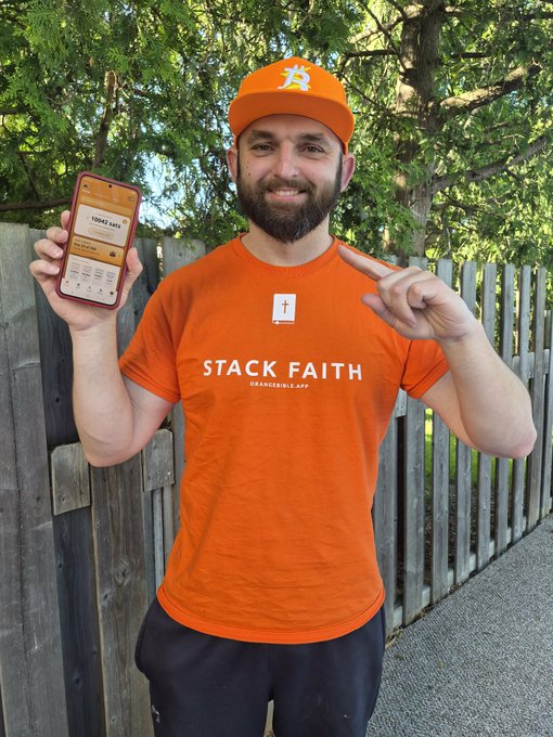The image features a man wearing an orange shirt with the words "Stack Faith" printed on it, holding up his phone. The man appears to be smiling and posing for the camera while showing off his phone screen. The background of the photo includes a wooden fence and trees, creating a pleasant outdoor setting.